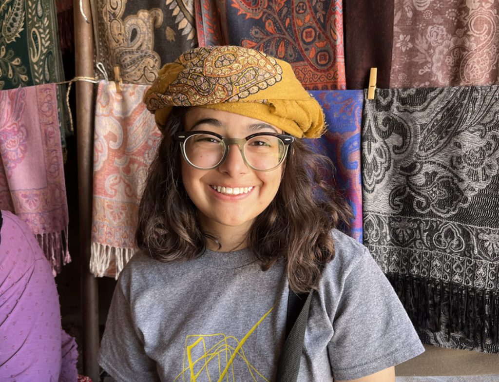 Reem smiles with a yellow headscarf, standing in front of several multicolored cloths.