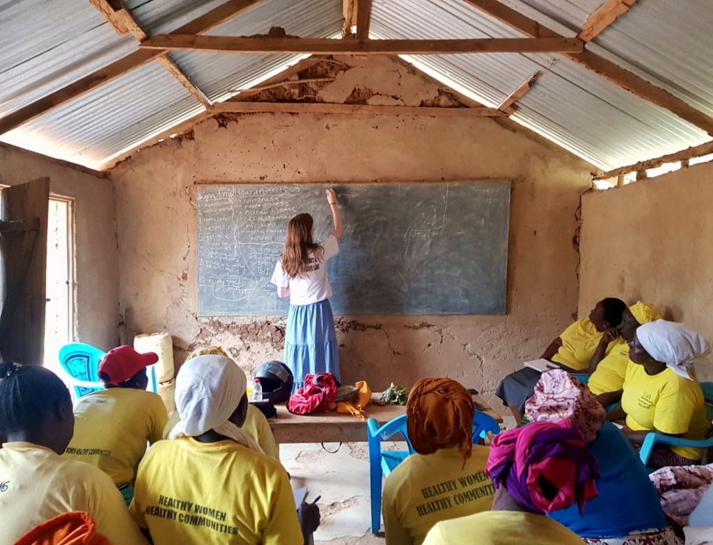 Jess stands with her back to a classroom full of people in yellow shirts, writing on a chalkboard.