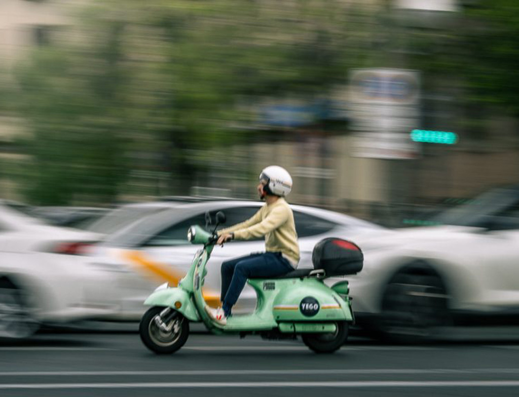 A mint green motorcycle is in focus along a street, passing by blurry cars in the background.