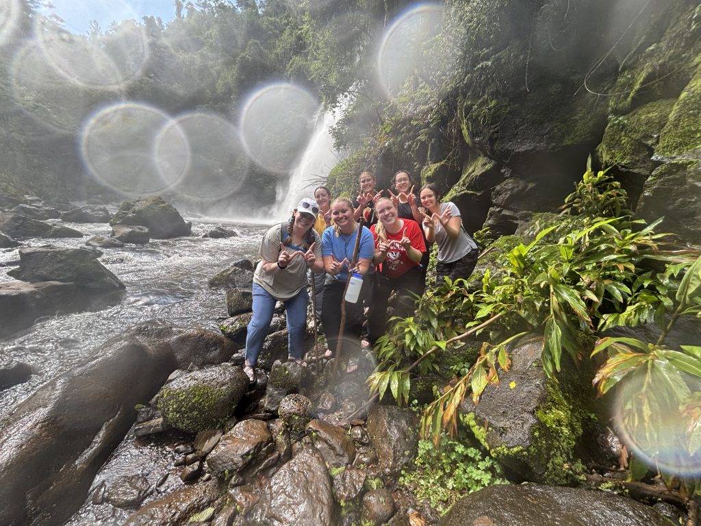 A group of people making a W with their fingers are standing in front of a waterfall, surrounded by tree branches and luscious green plants.