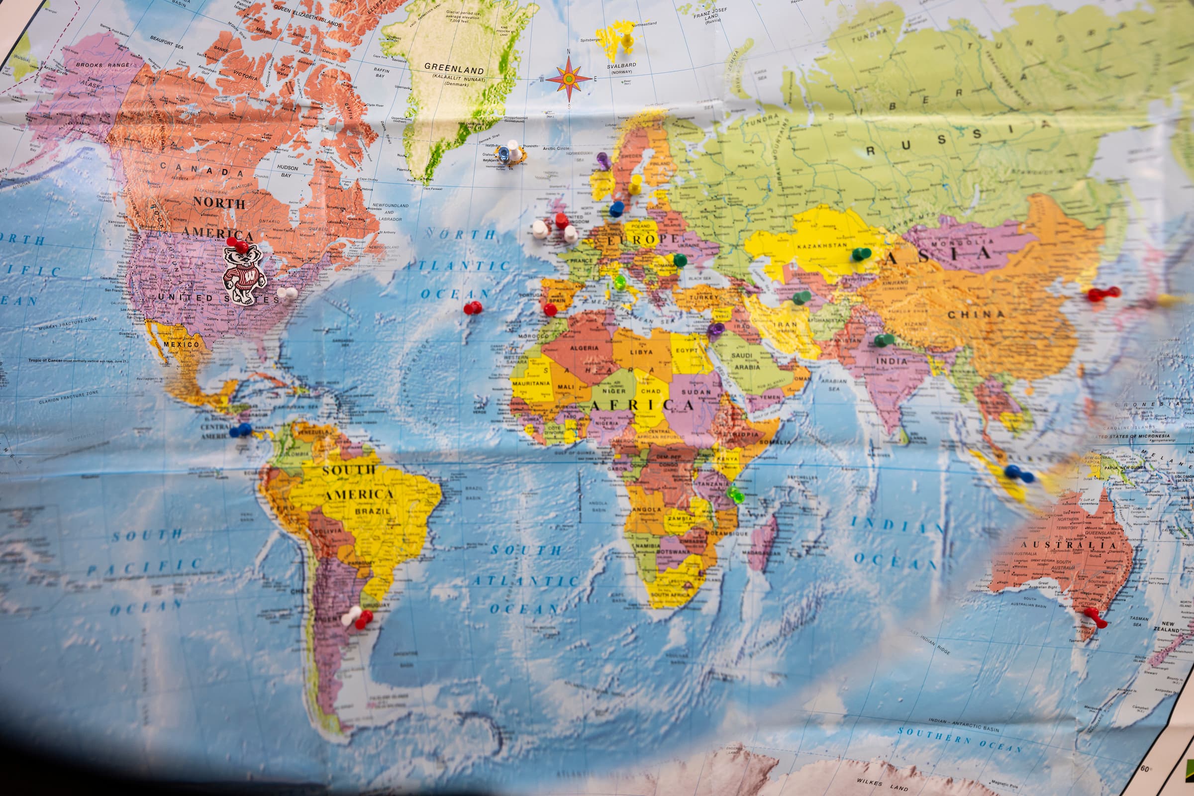 Colorful world map with push pins at different visited locations across the glob including a picture of Bucky Badger in Wisconsin.